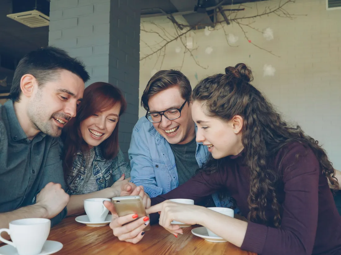 カフェでスマートフォンを囲んで笑う4人の友人